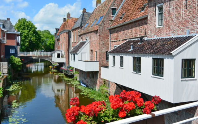 Canal lined with brick buildings, red flowers in the foreground, and a bridge in the background under a blue sky with clouds. "Hanging kitchens" in Appingedam, Netherlands