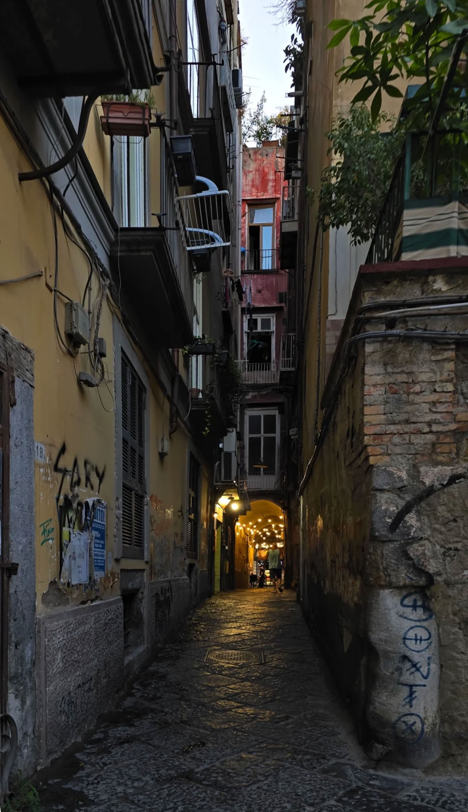 Narrow cobblestone alleyway between tall buildings with balconies, graffiti on walls, and a lit doorway at the end.