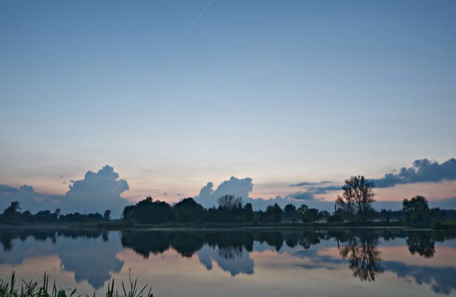 A serene lake reflecting the sky and clouds at sunset, with silhouettes of trees along the horizon.