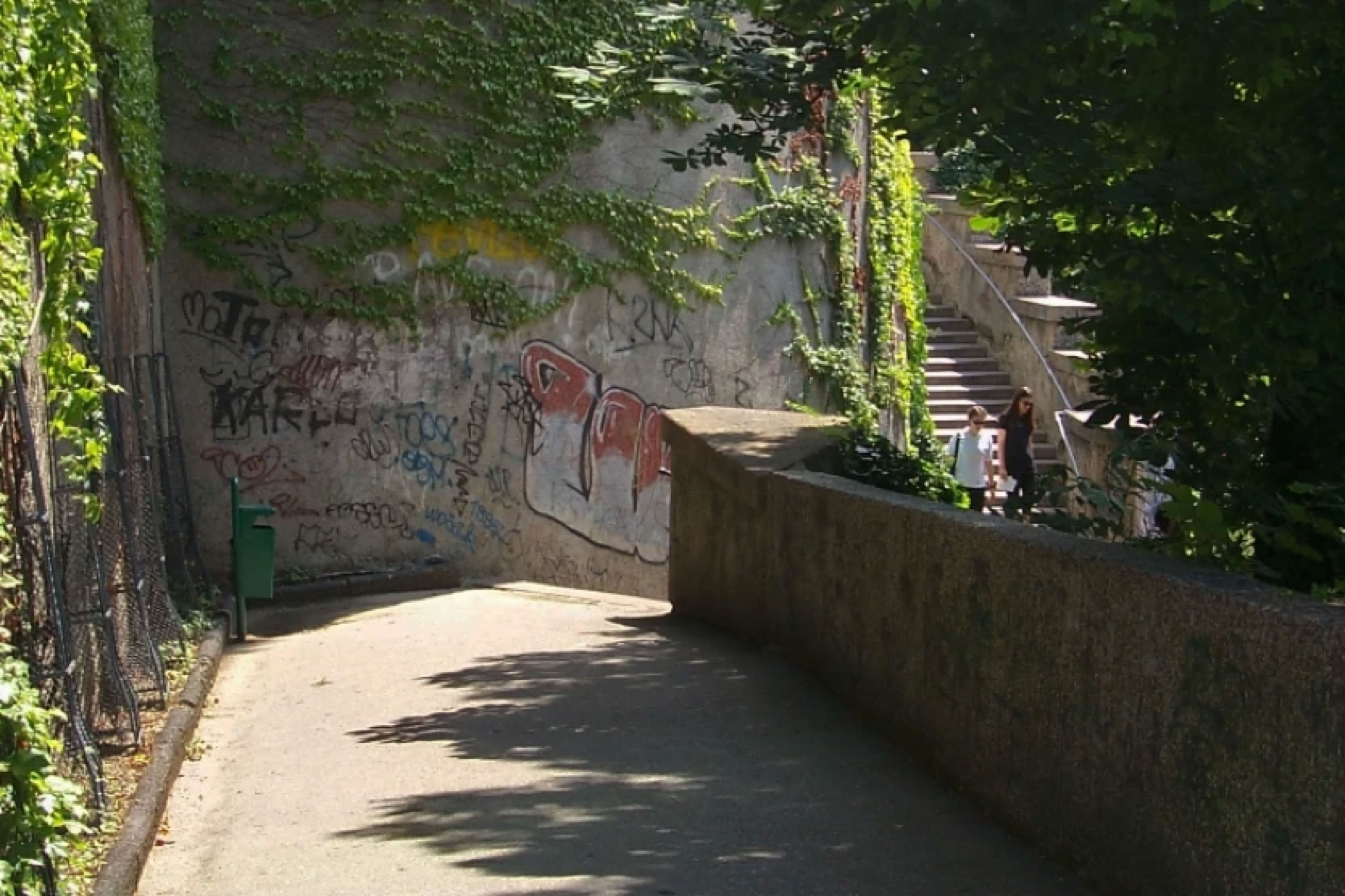 A narrow pathway with a stone wall on the right, covered in graffiti. The path is lined with greenery and leads to a set of stairs in the background.