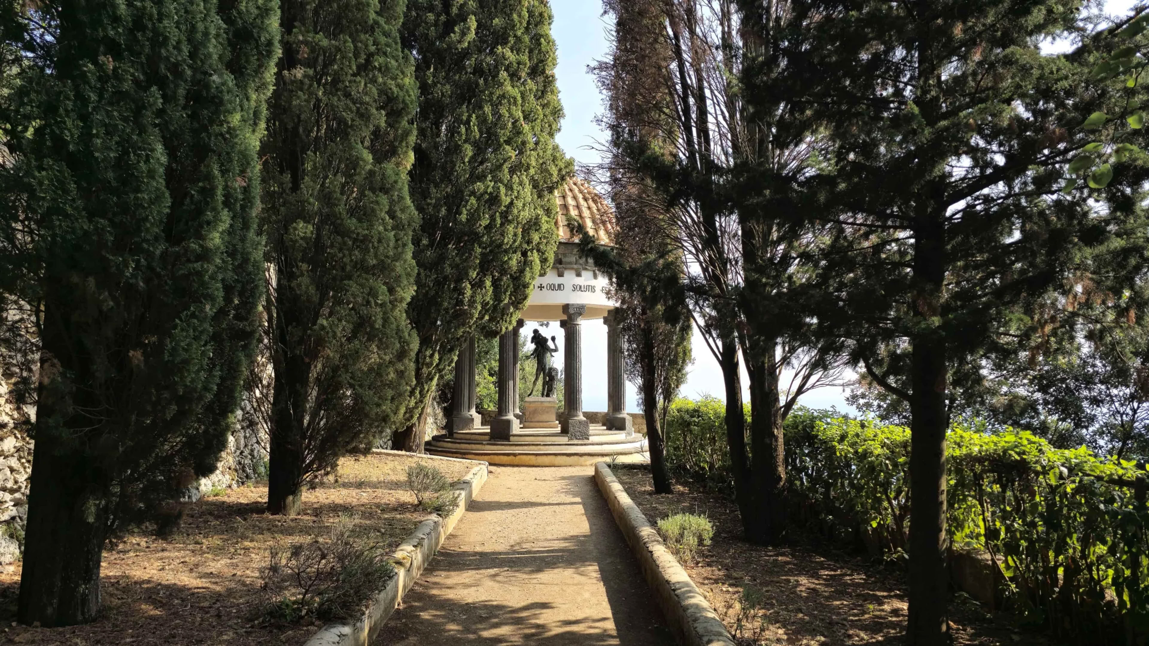 A tree-lined pathway leading to a small structure with columns, surrounded by greenery.