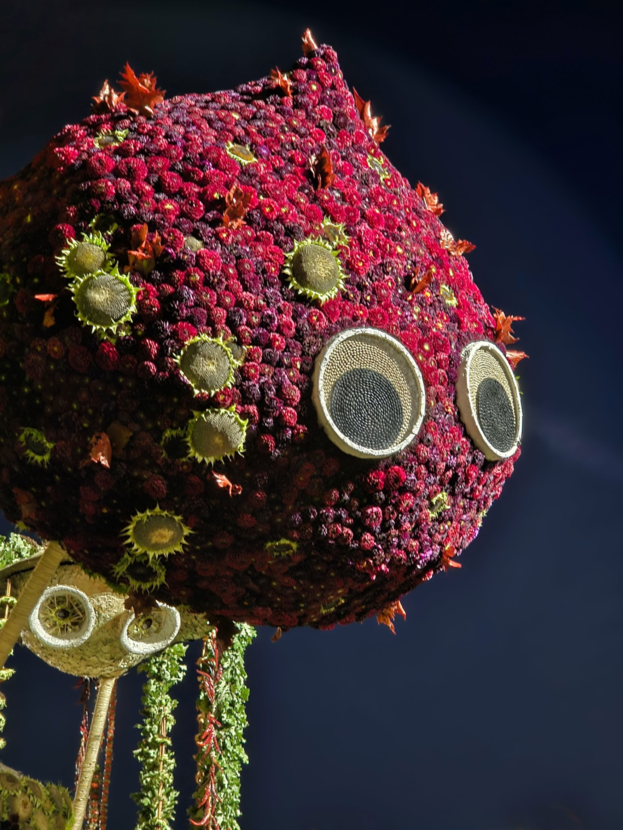 A large, spherical structure covered in red flowers with circular, eye-like features and green floral accents, set against a dark background.