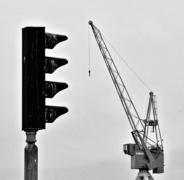 A black and white image showing a traffic light on the left and a large crane on the right against a plain sky.