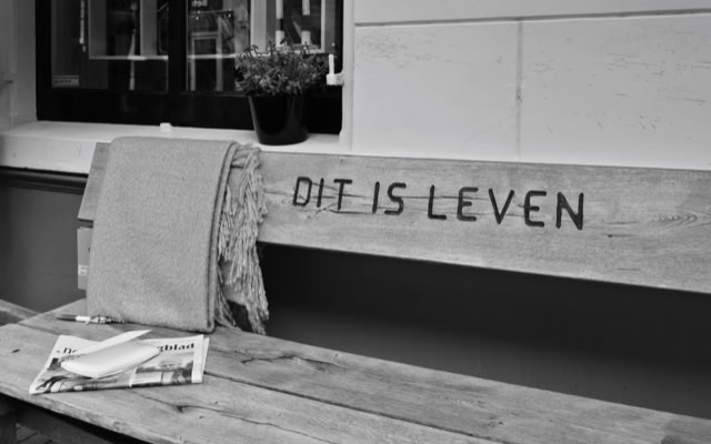 A wooden bench with a blanket draped over one side and a newspaper on the seat. The words "DIT IS LEVEN" are carved into the backrest. A small plant is visible in the background.
