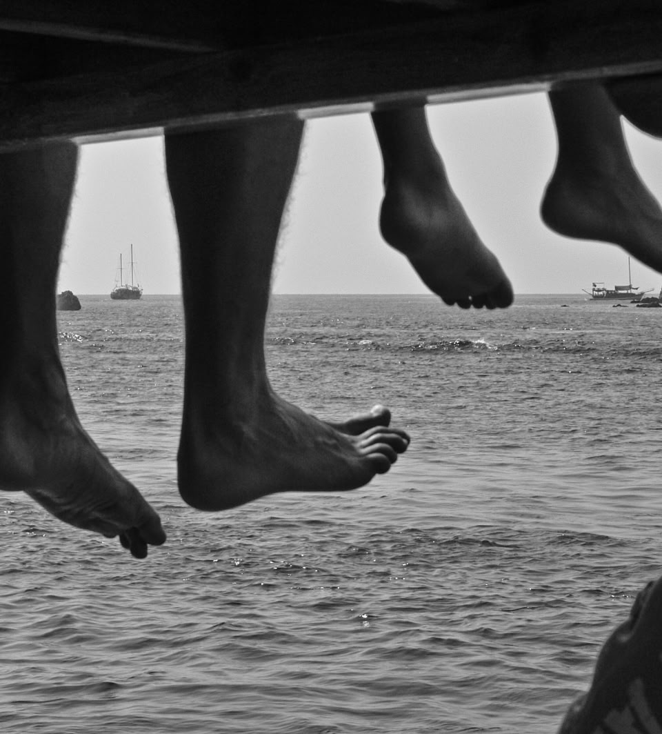 Three pairs of legs dangling from a structure above water, with boats visible in the distance.