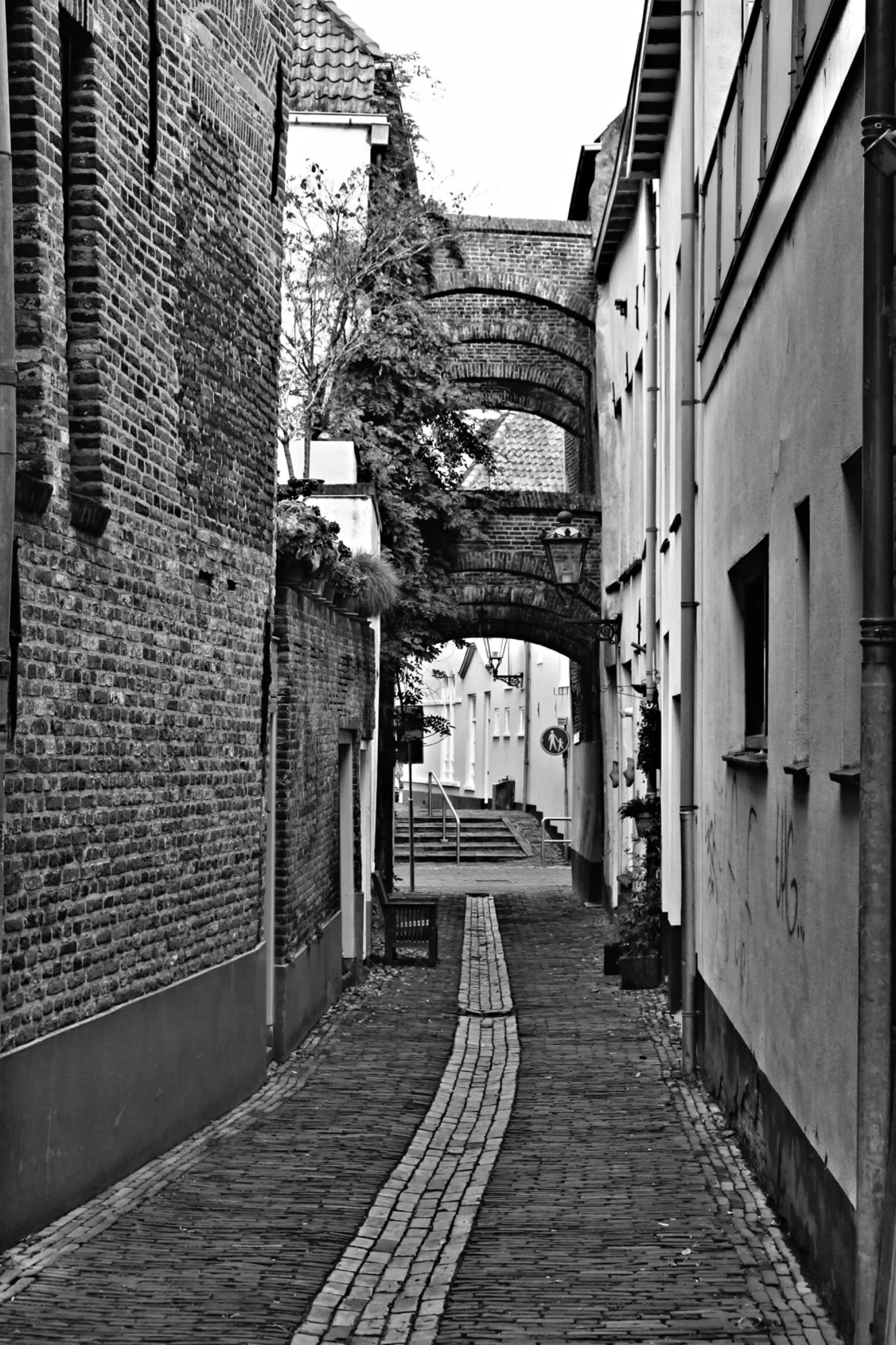 Narrow cobblestone alleyway between brick and plaster buildings, with an arched bridge overhead.