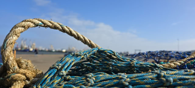 Close-up of a pile of blue fishing nets with a thick rope on a beach, with a clear blue sky and distant buildings in the background.