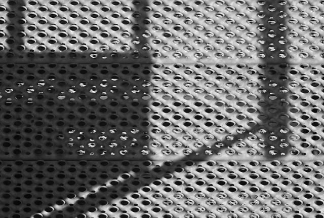 A black and white image showing a perforated surface with circular holes, casting geometric shadows.