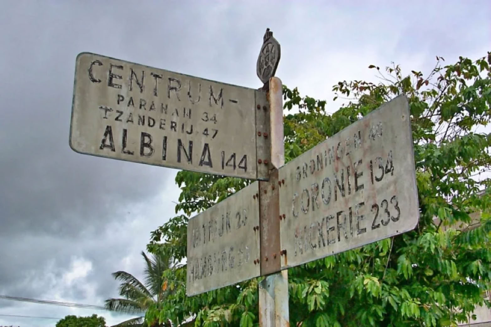 A weathered street sign with multiple directional arrows and text, set against a backdrop of cloudy sky and green foliage.
