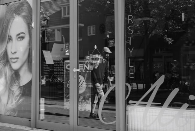 A black and white image of a storefront with large windows, displaying a poster of a woman with long hair on the left. Reflections of trees and buildings are visible in the glass.