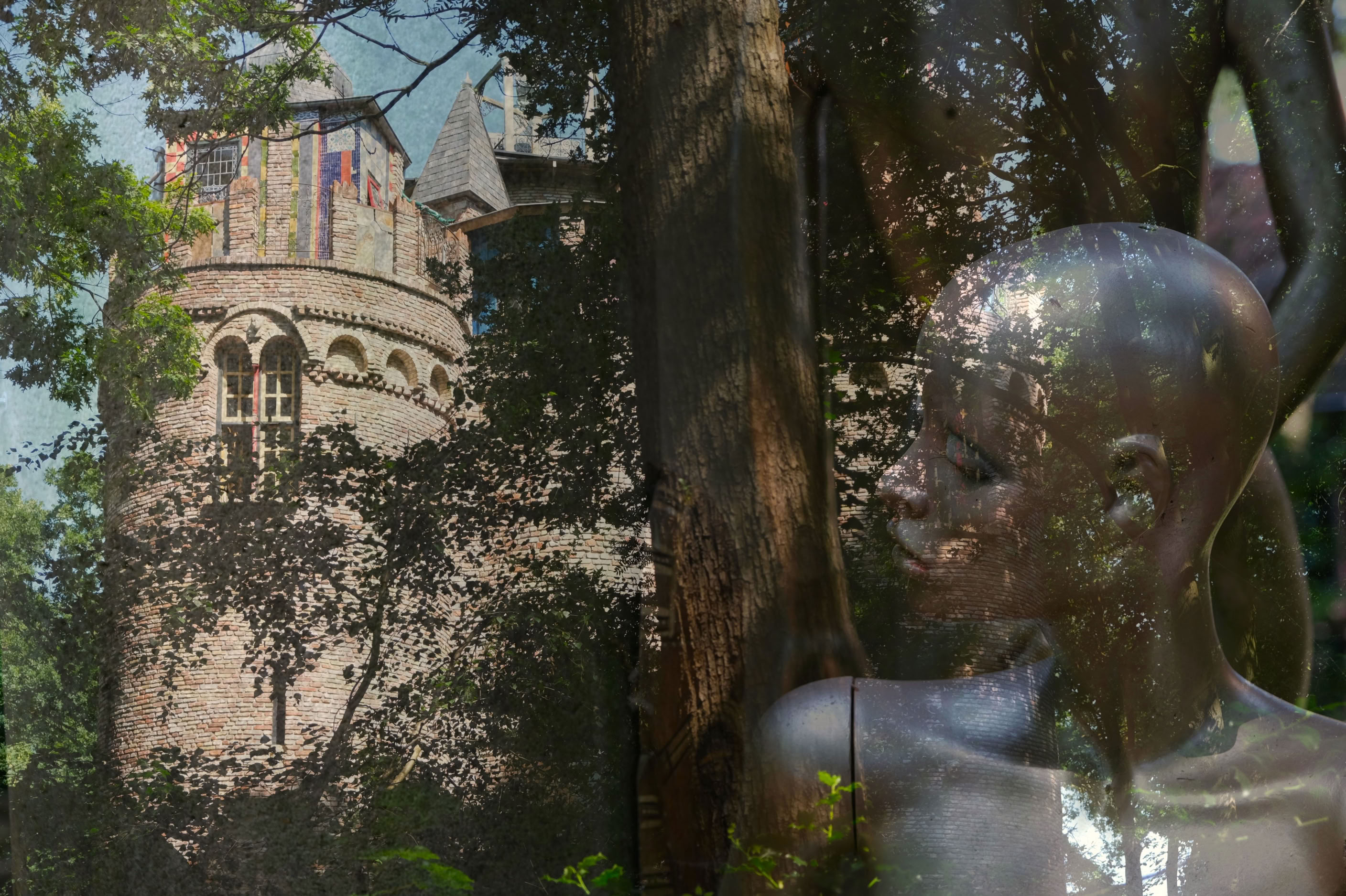 A stone tower partially obscured by trees is overlaid with a reflective surface showing a sculpture of a human figure.