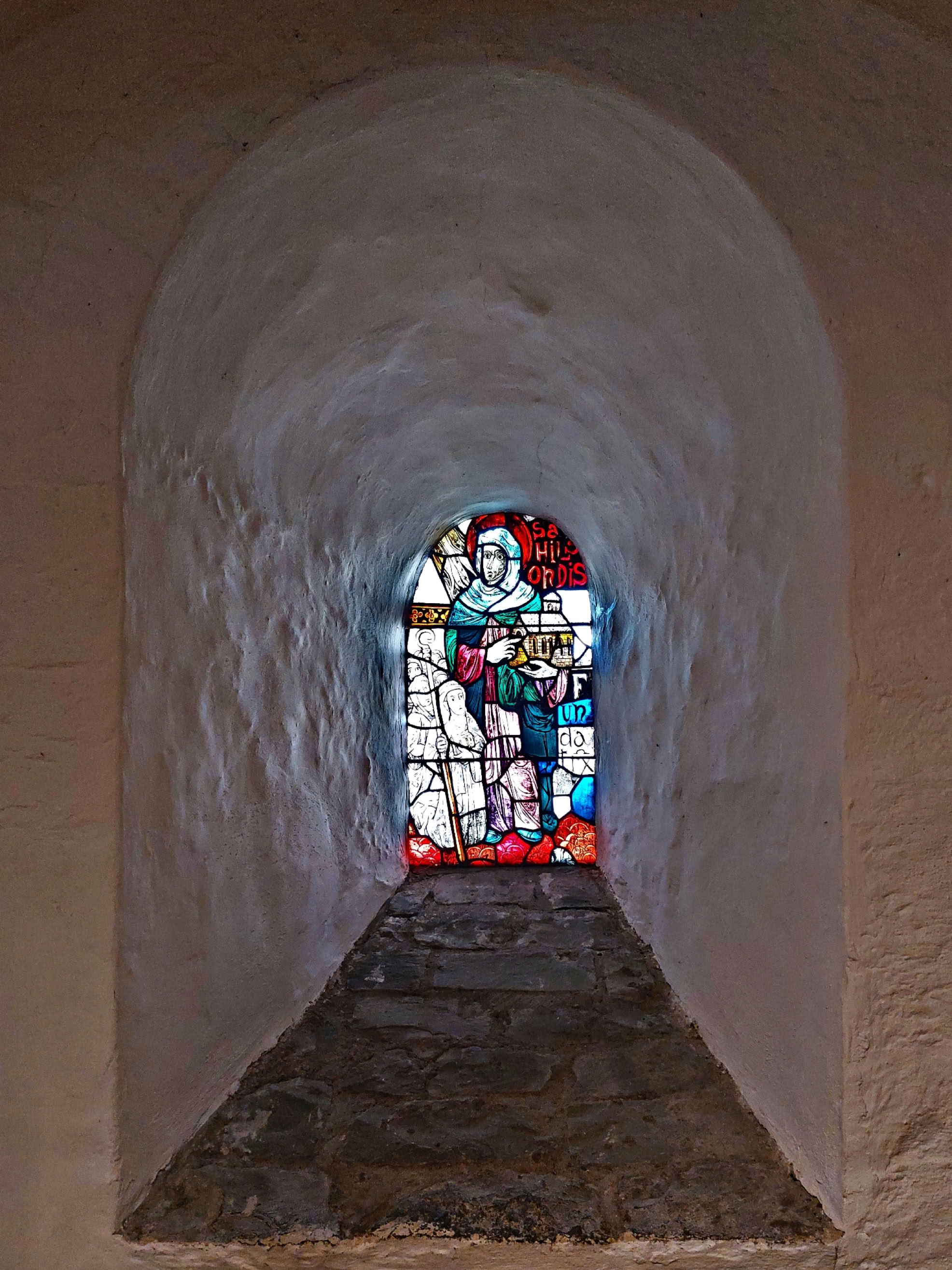 A narrow stone corridor leading to a colorful stained glass window depicting figures.