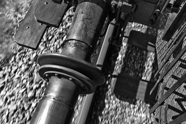 Black and white image of a train wheel and axle on a track with gravel beneath.