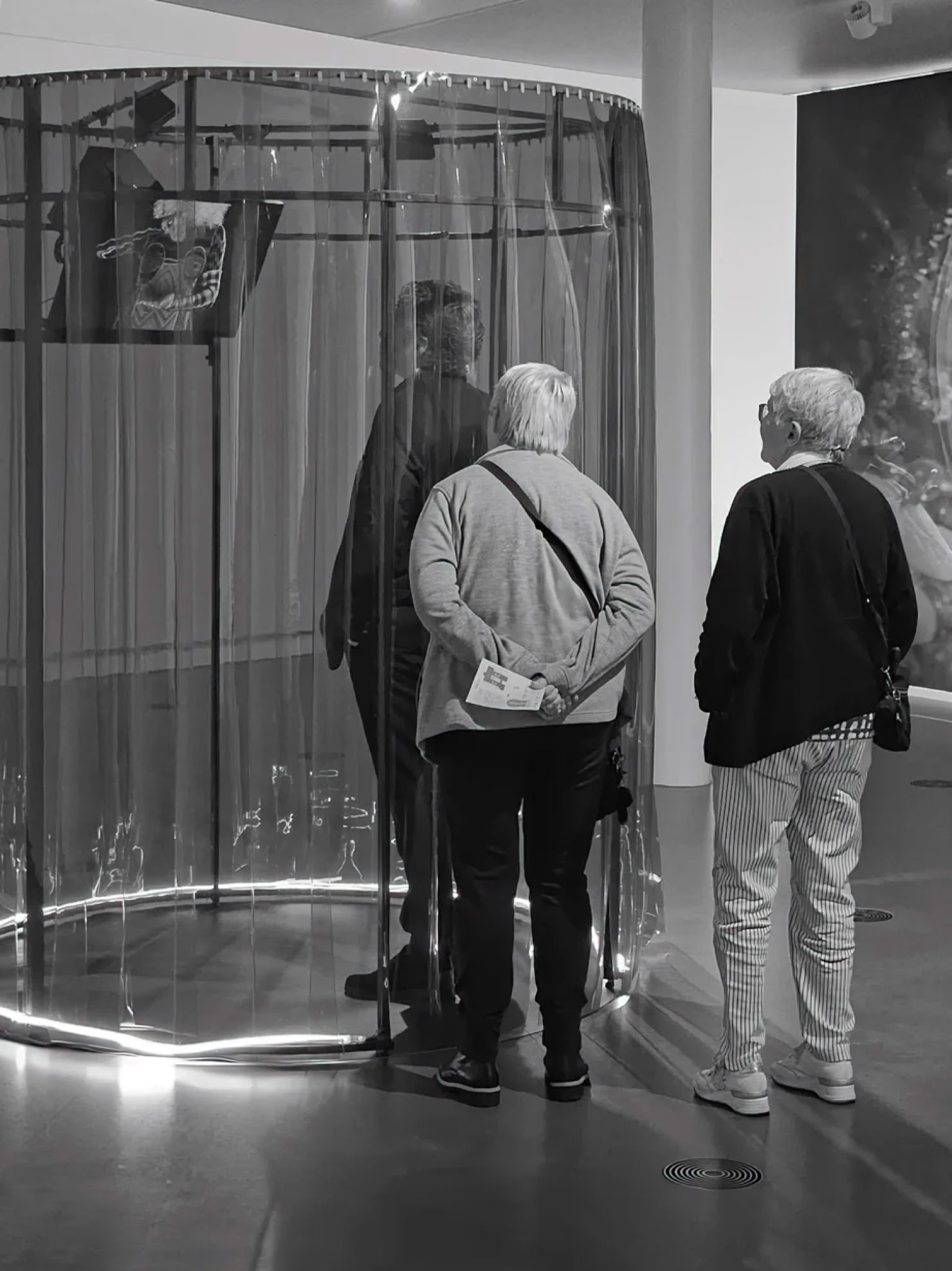 Two people standing in front of a circular installation with transparent curtains, viewing an exhibit inside a gallery.
