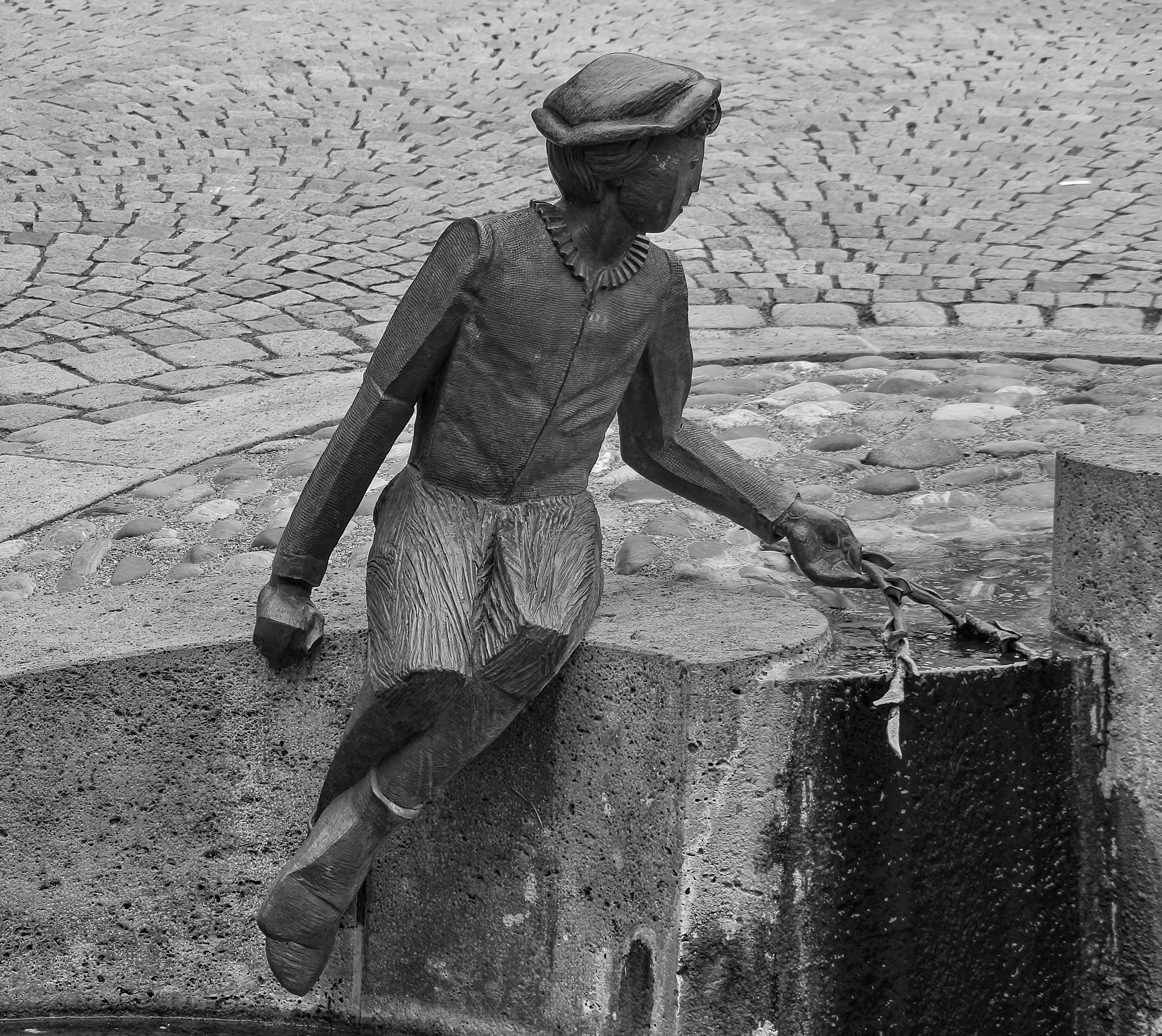 A statue of a person sitting on a stone ledge, holding a chain, with a cobblestone ground in the background.