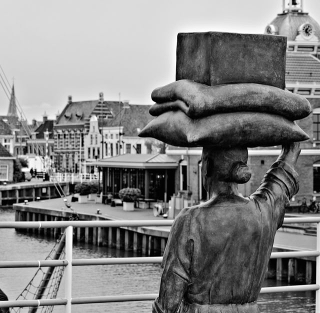 A black and white image of a statue depicting a person carrying a stack of bags or parcels on their head, with a cityscape and waterway in the background.
