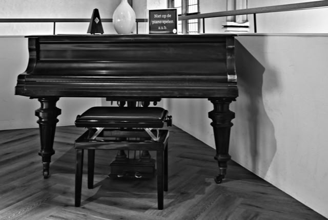 A black and white image of a grand piano with a bench in front, placed on a wooden floor. A vase and the text "Please do not play on this piano" on top of the piano.