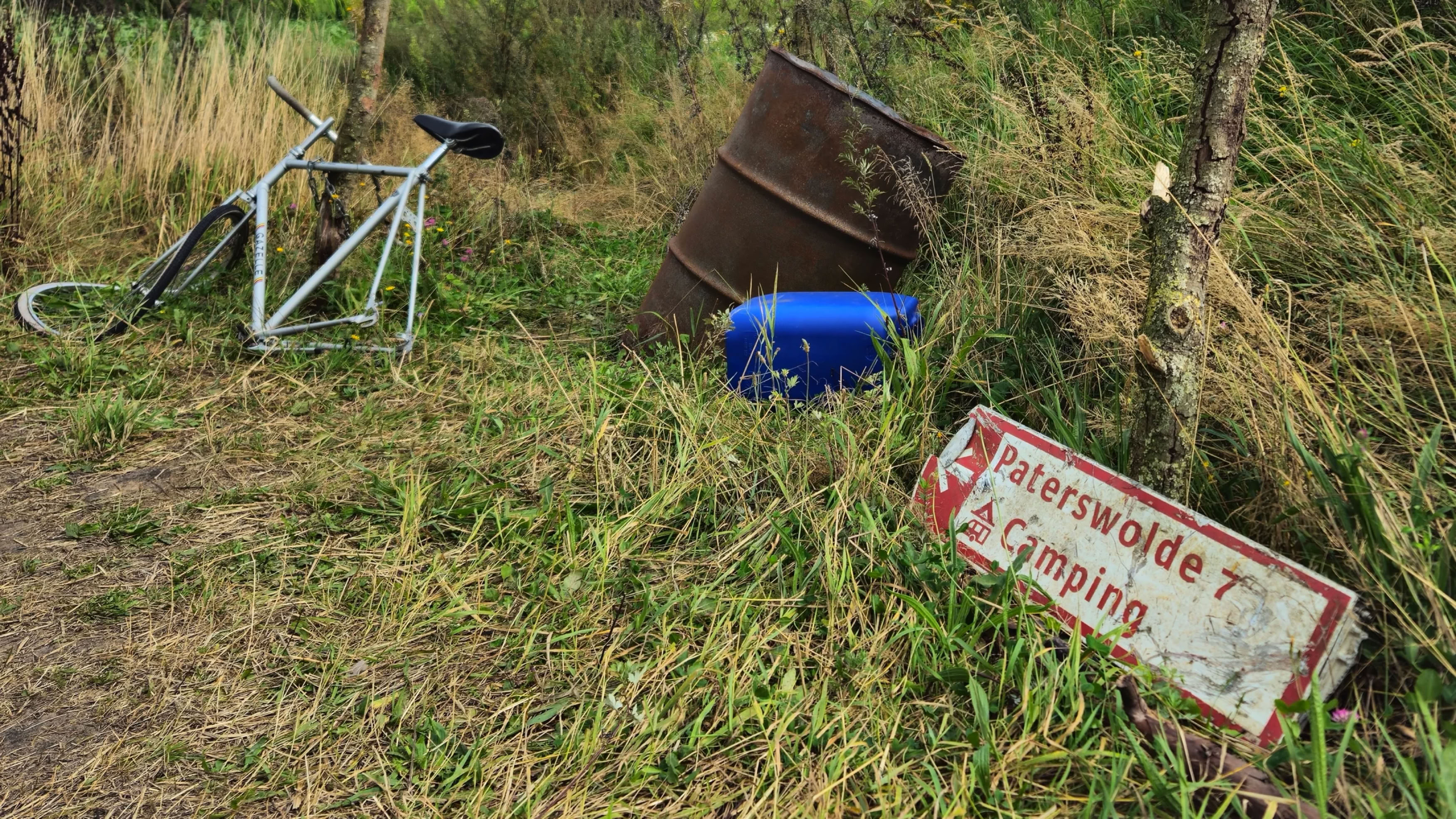 A bicycle, a rusted barrel, a blue container, and a tilted sign with text are on grassy ground.