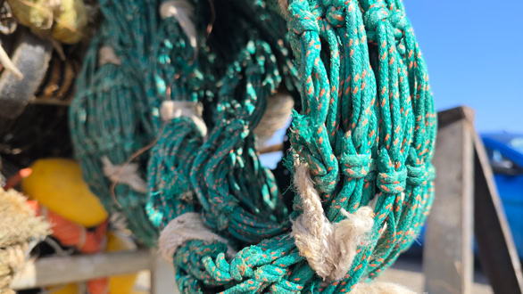 Close-up of a green fishing net with hooks, against a blurred background of a blue sky and part of a boat.
