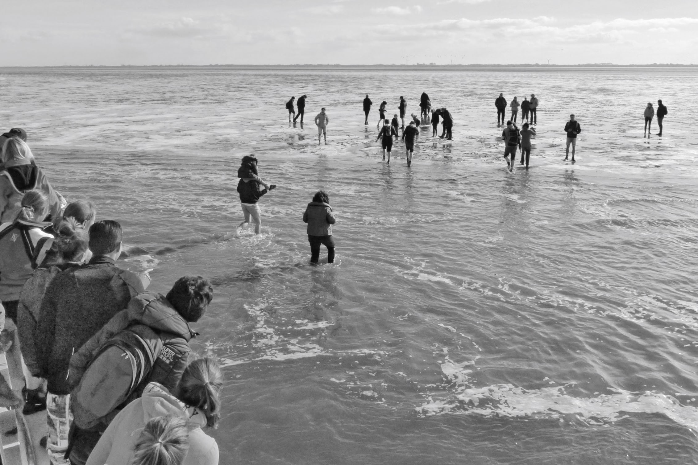 A group of people standing and wading in shallow water, with some onlookers on the left side of the image.