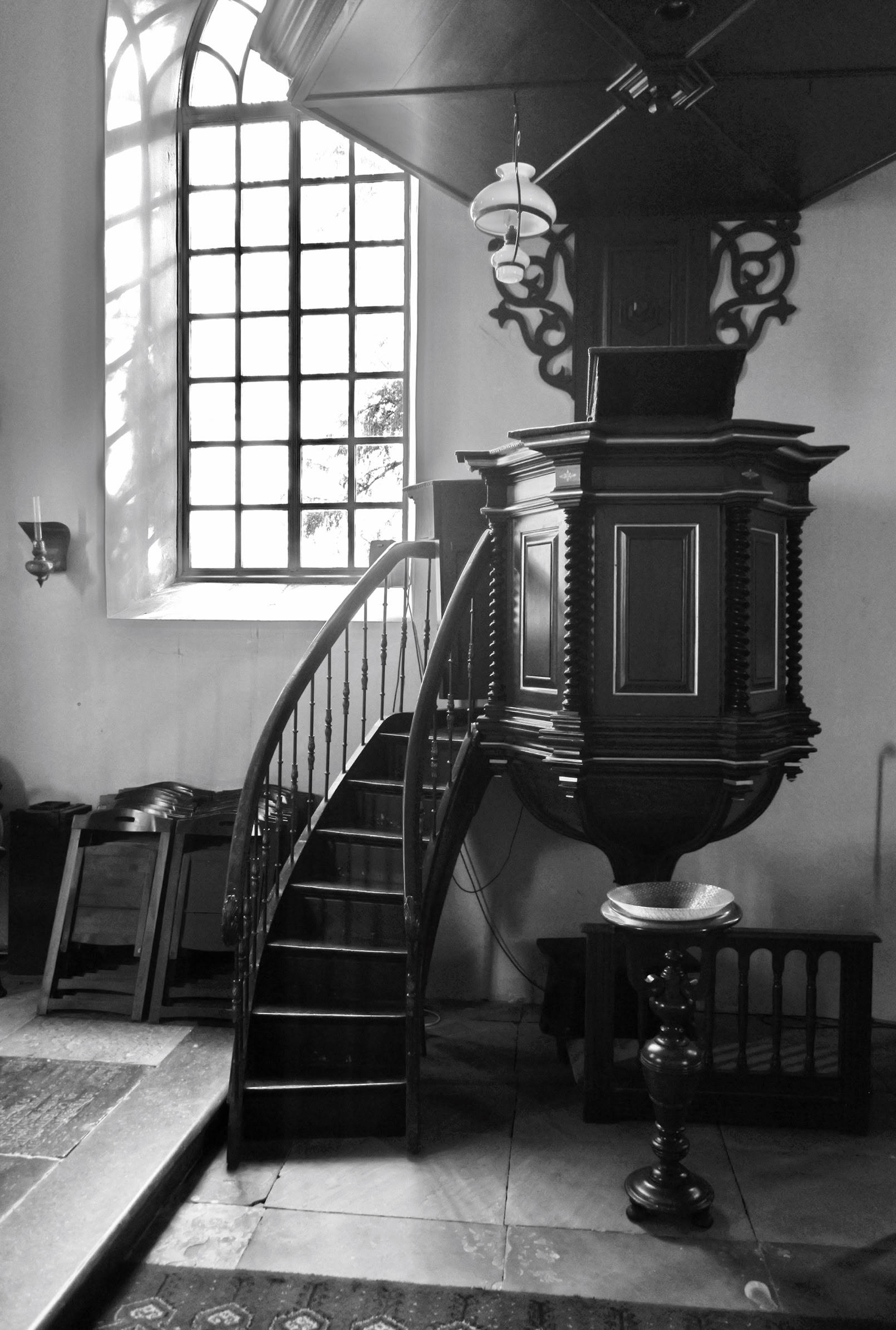 A black and white image of an ornate wooden pulpit with a staircase, situated in a church  with a large window.