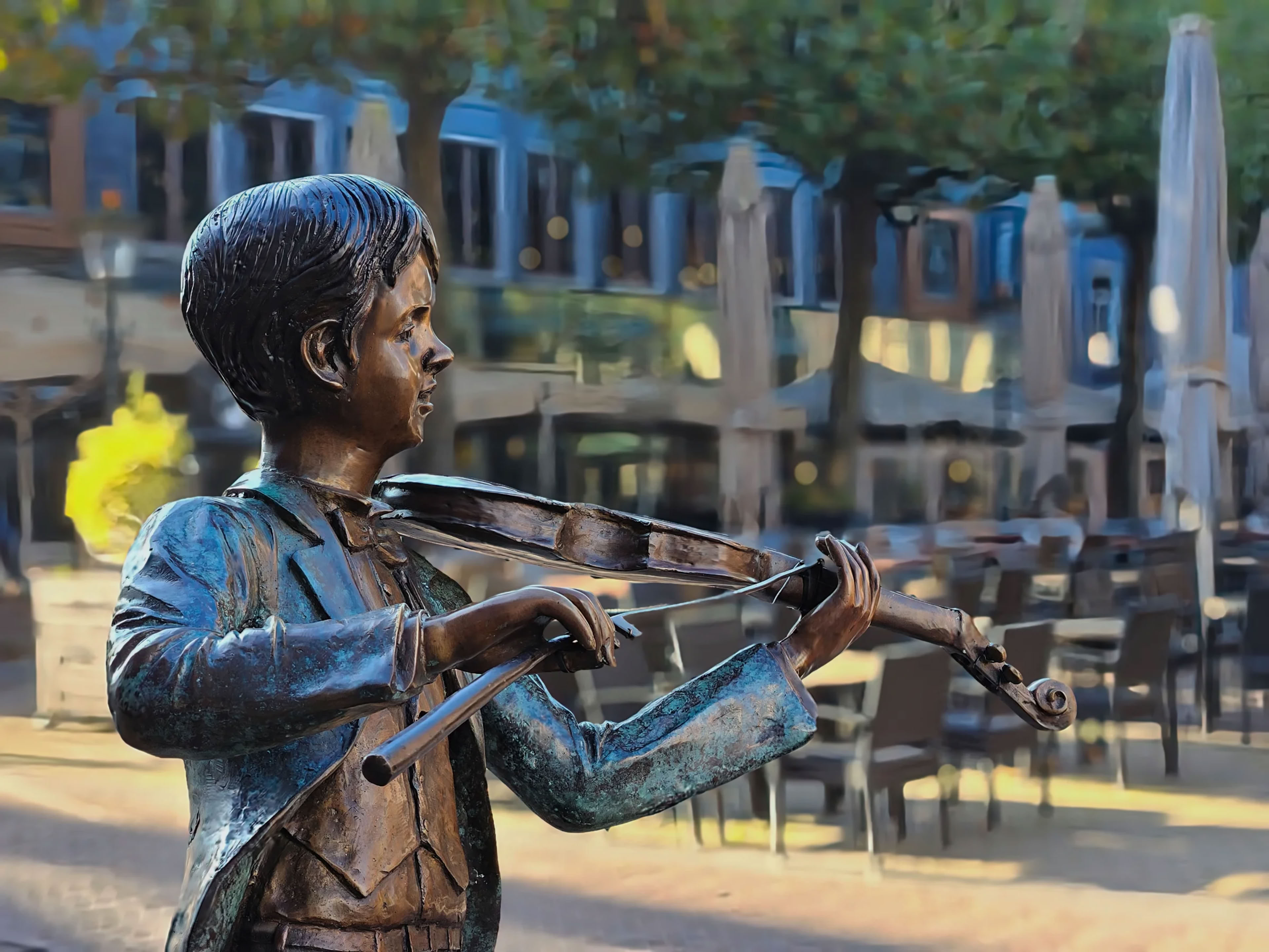 A bronze statue of a child playing the violin, set in an outdoor cafe area with tables, chairs, and closed umbrellas in the background.