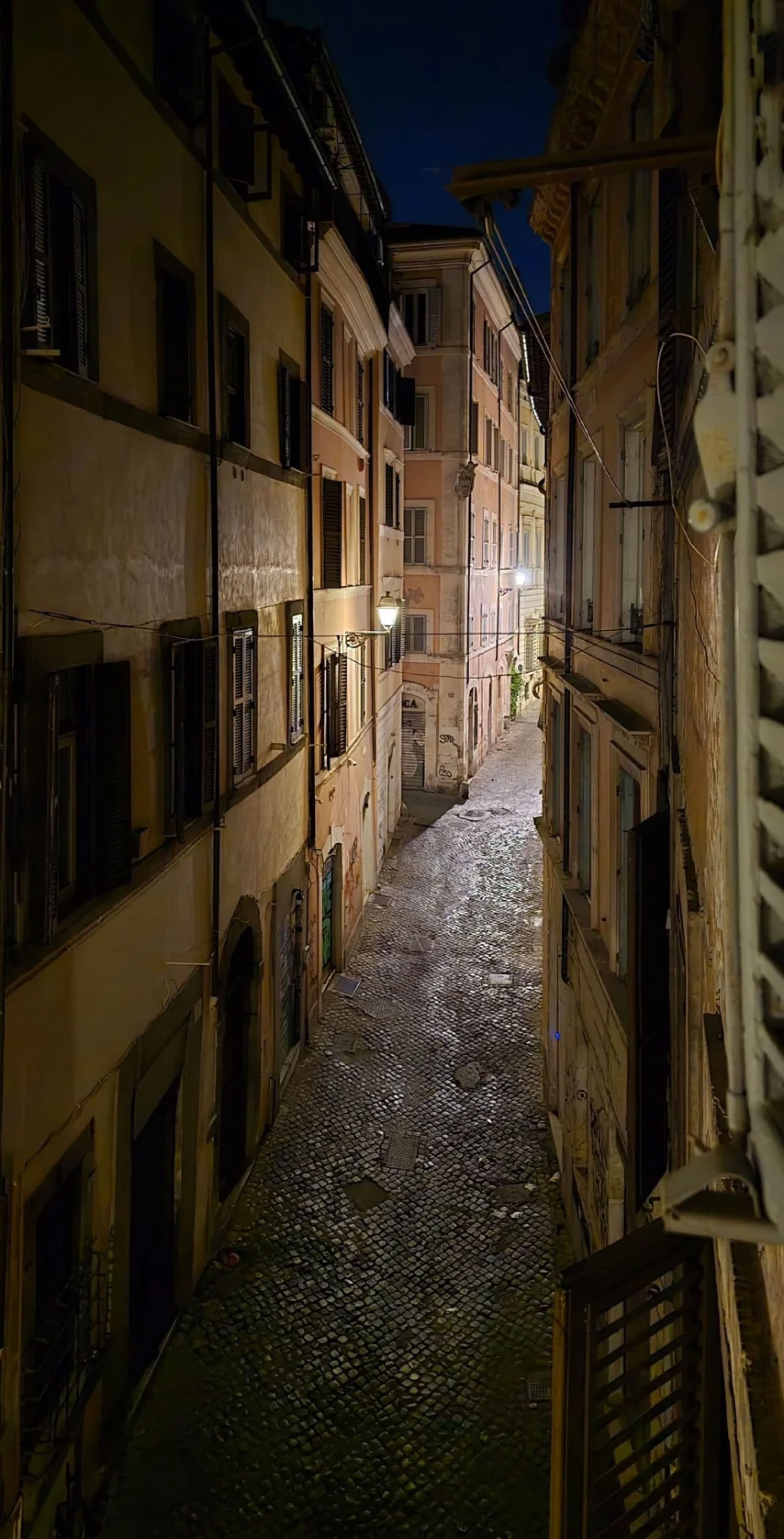Narrow cobblestone alleyway between tall, closely spaced buildings at night, with dim street lighting.