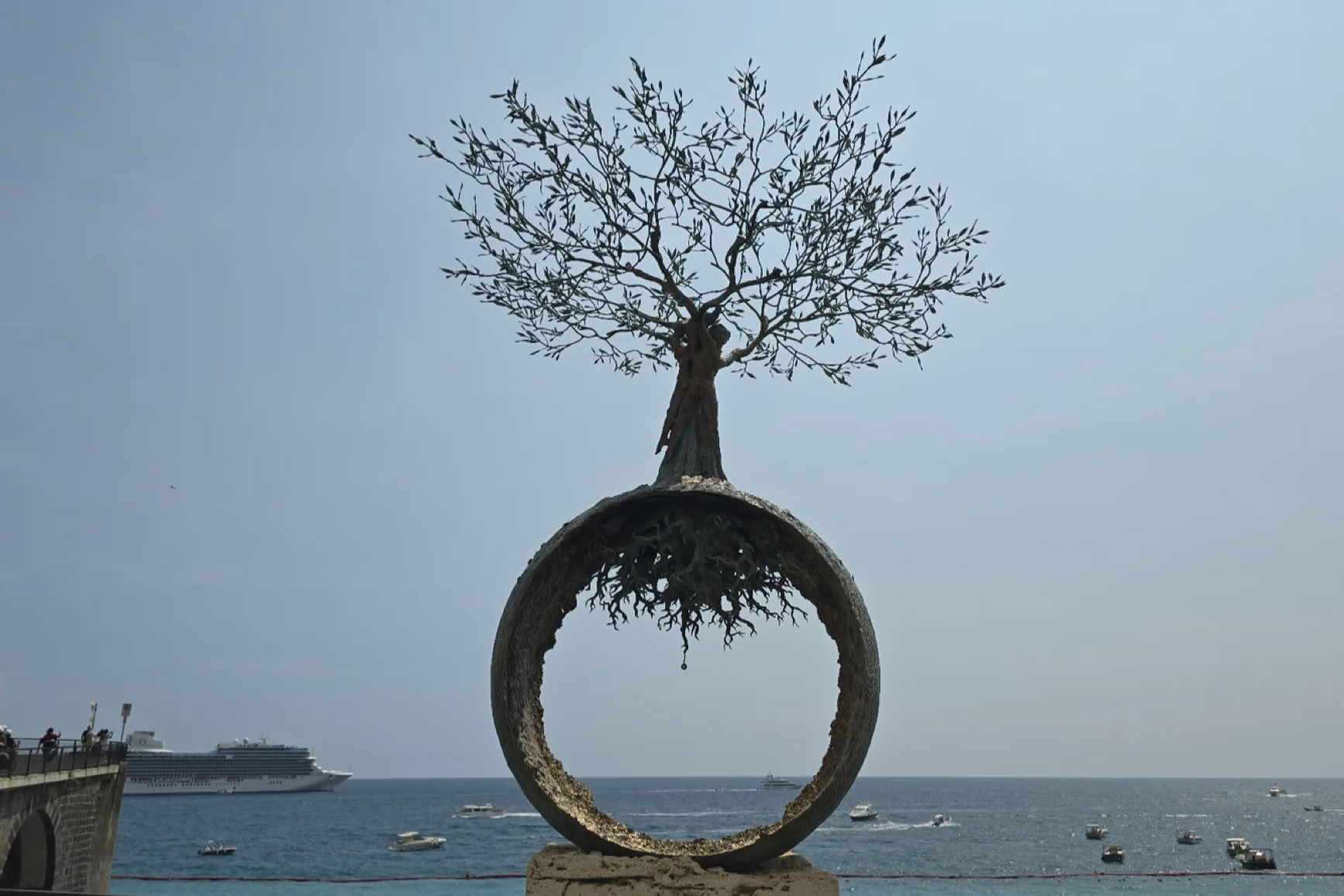 A tree growing on top of a large circular sculpture with roots visible, set against a backdrop of the sea and a clear sky.