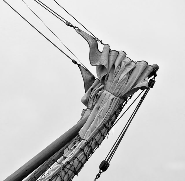 Close-up of a ship's bow with rigging and a furled sail against a cloudy sky.